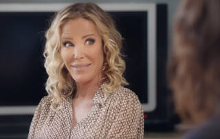 Femme aux cheveux blonds bouclés portant une chemise à motifs dans un environnement neutre avec un écran de télévision.