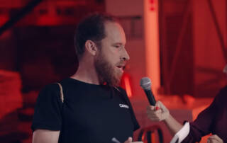 Image d'une interview avec un homme barbu portant un t-shirt noir 'CANAL' et une femme tenant un microphone sous des lumiè...