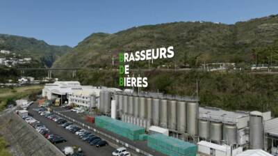 Aérophoto d'une usine avec des silos métalliques, des conteneurs verts et un parking. Texte 'BRASSEURS DE BIÈRES' en surim...