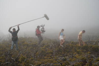 Deux techniciens et une actrice en tenue blanche sur un plateau de tournage en forêt avec brume.