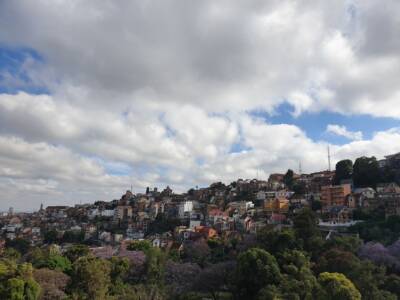 Panorama de ville sur colline, bâtiments multicolores avec toits rouges et blancs, arbres et fleurs en avant-plan, ciel nu...