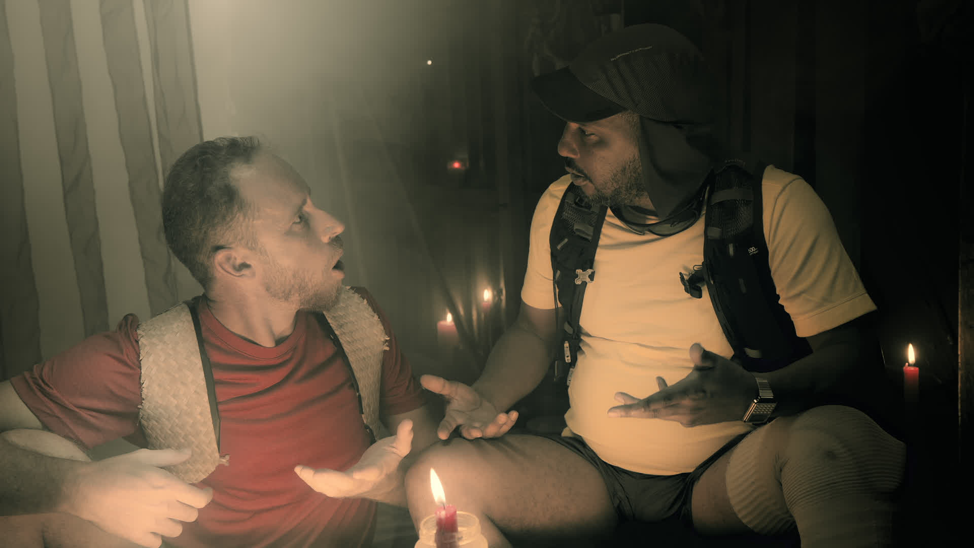 Deux hommes en discussion dans une pièce sombre éclairée par des bougies, l'homme à gauche porte un gilet rouge et un t-sh...