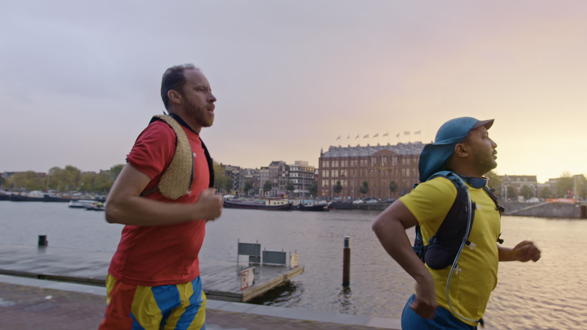 Deux personnes en short et t-shirt courent le long d'un quai avec des bâtiments, des arbres et des bateaux amarrés. Ciel c...