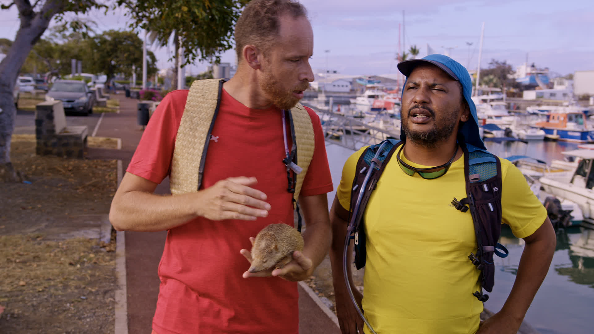 Deux hommes en plein air près d'un port avec des bateaux amarrés, l'homme gauche tient un animal, l'autre porte une casque...