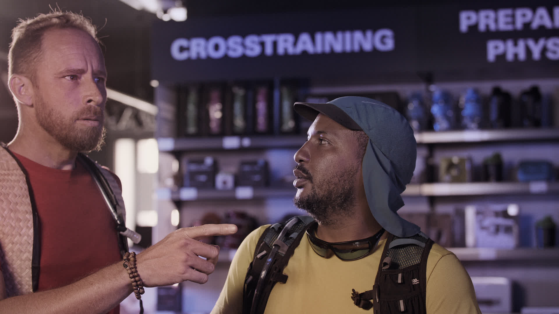 Deux hommes portant des vêtements colorés discutent près d'étagères avec un écran affichant 'CROSSTRAINING' dans un intéri...
